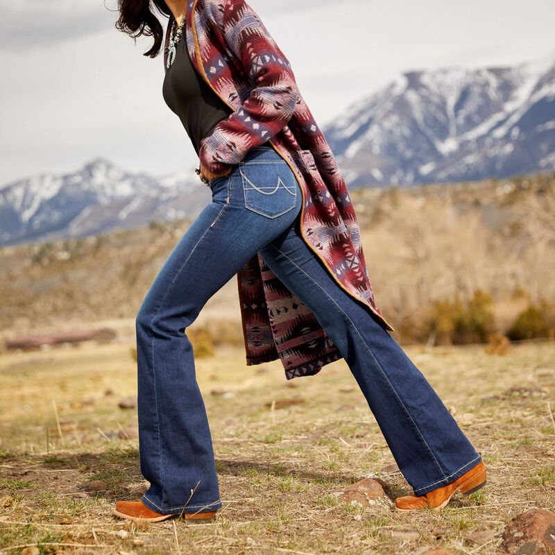 Person wearing blue jeans and a patterned shirt in a mountainous landscape