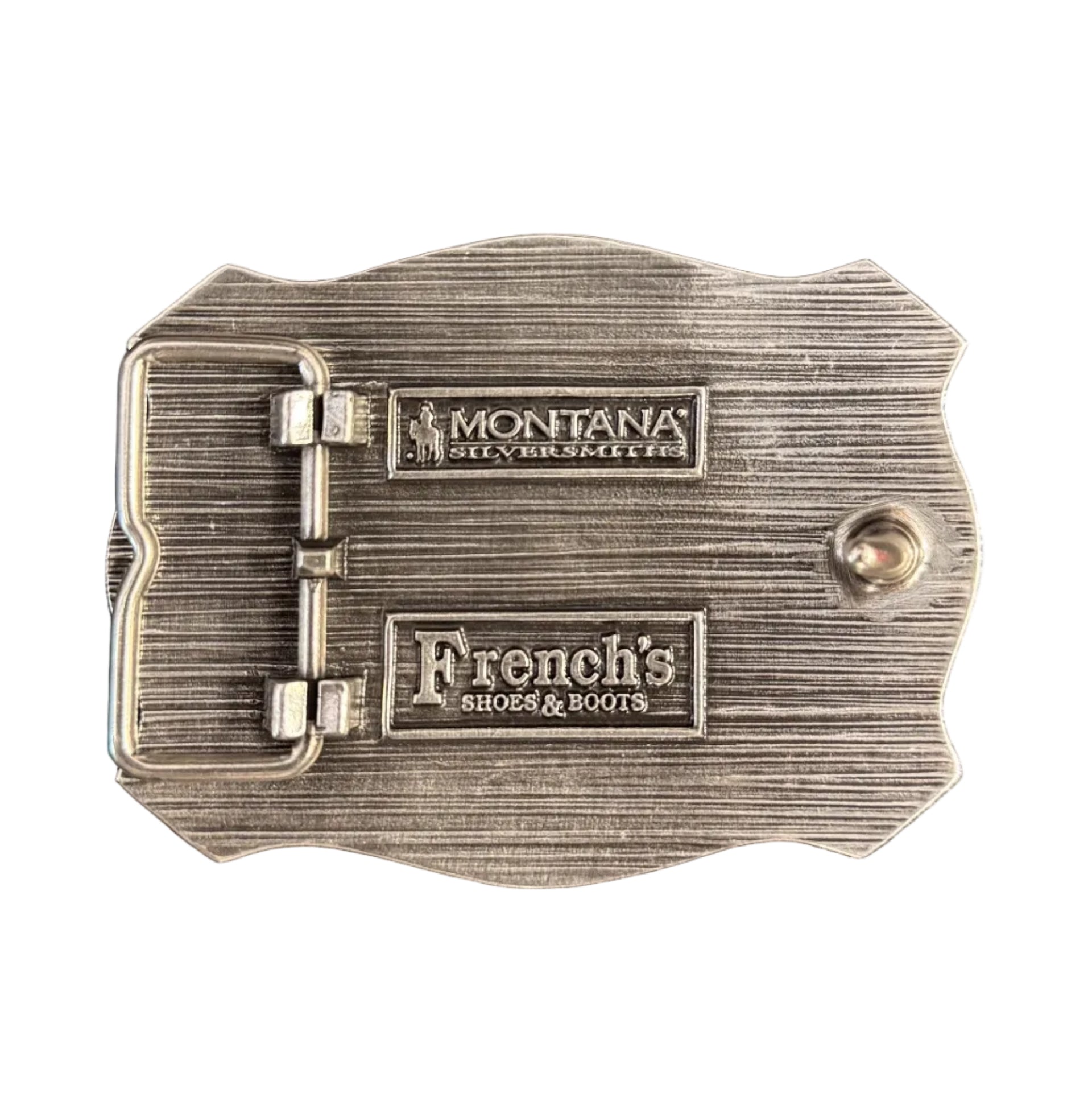 A silver-toned metal buckle with a textured brushed finish, featuring two engraved rectangular plates that read 'Montana' and 'French's' with smaller text below, photographed on a dark surface.