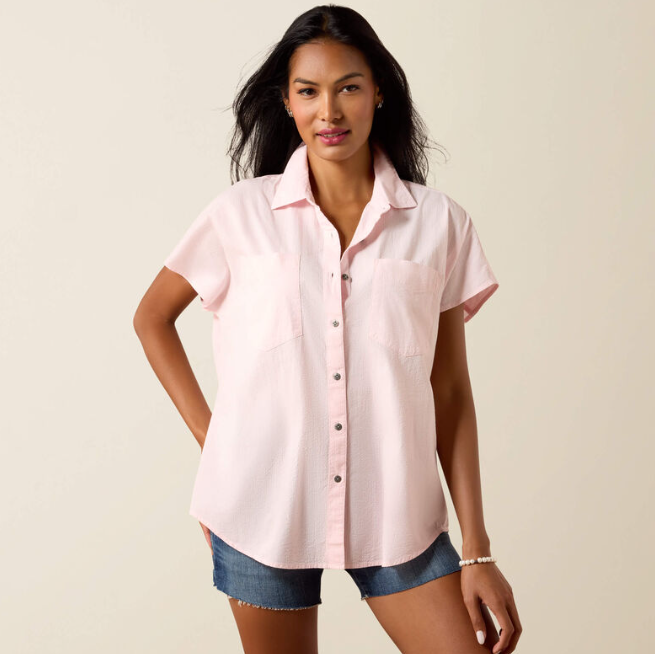 A woman modeling a light pink short-sleeve button-up shirt with a chest pocket, paired with denim shorts, against a plain beige studio background.