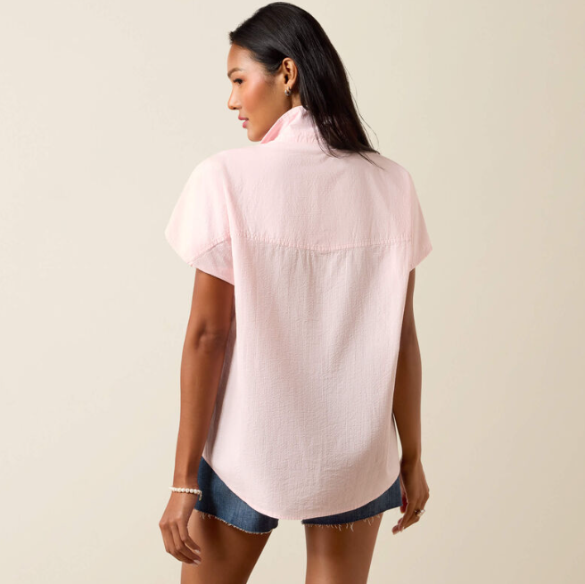 A woman shown from the back wearing a light pink short-sleeve button-up shirt and denim shorts against a plain beige studio background.