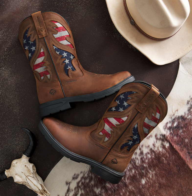 Brown cowboy boots with American flag design on a rustic surface with a hat and cow skull.
