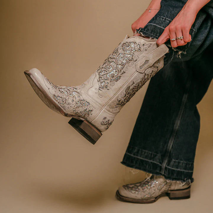 Person wearing white cowboy boots with intricate designs on a beige background