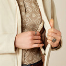 Close-up of hands wearing rings, holding a beige jacket with a patterned shirt underneath.