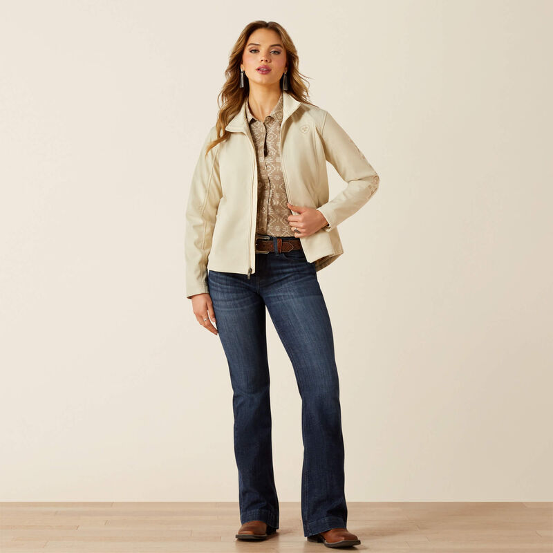 Woman wearing a beige jacket, patterned shirt, and blue jeans on a plain background