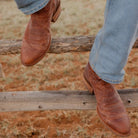 A close-up lifestyle shot of a person's legs dangling over a rustic wooden fence, wearing light blue jeans and brown leather western cowboy boots.
