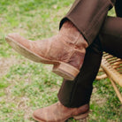 A close-up of a person's legs crossed, wearing a pair of distressed brown leather cowboy boots and dark brown trousers, sitting on a wicker chair outdoors on a grassy lawn.
