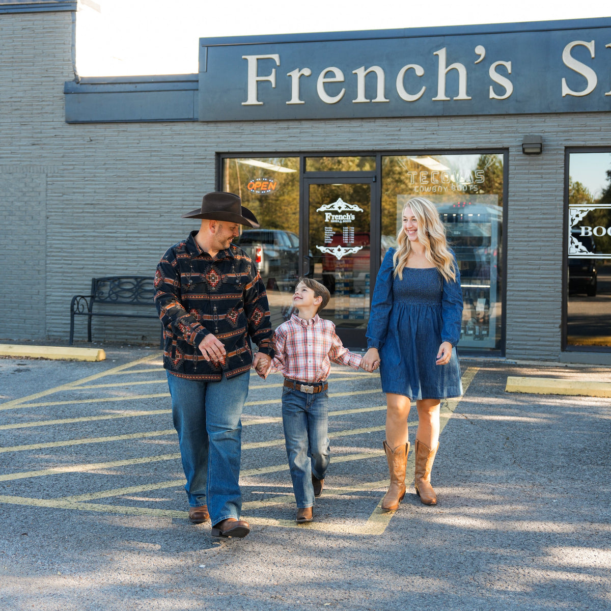 A family of three walks hand-in-hand across a paved parking lot in front of a brick storefront with a blue sign reading 'French's S.