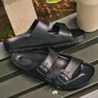 Black sandals on a wooden surface with a plant and container in the background