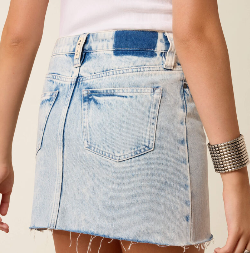 A close-up of a model wearing a light-wash blue denim mini skirt with a frayed hem, styled with a white ribbed top and a chunky silver cuff bracelet.