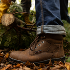 Brown work boot worn by a person in a forest setting with a log and foliage.