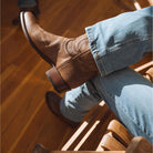 A person sitting on a wooden chair wearing a brown leather cowboy boot with western stitching, paired with light blue jeans, on a warm wood floor.
