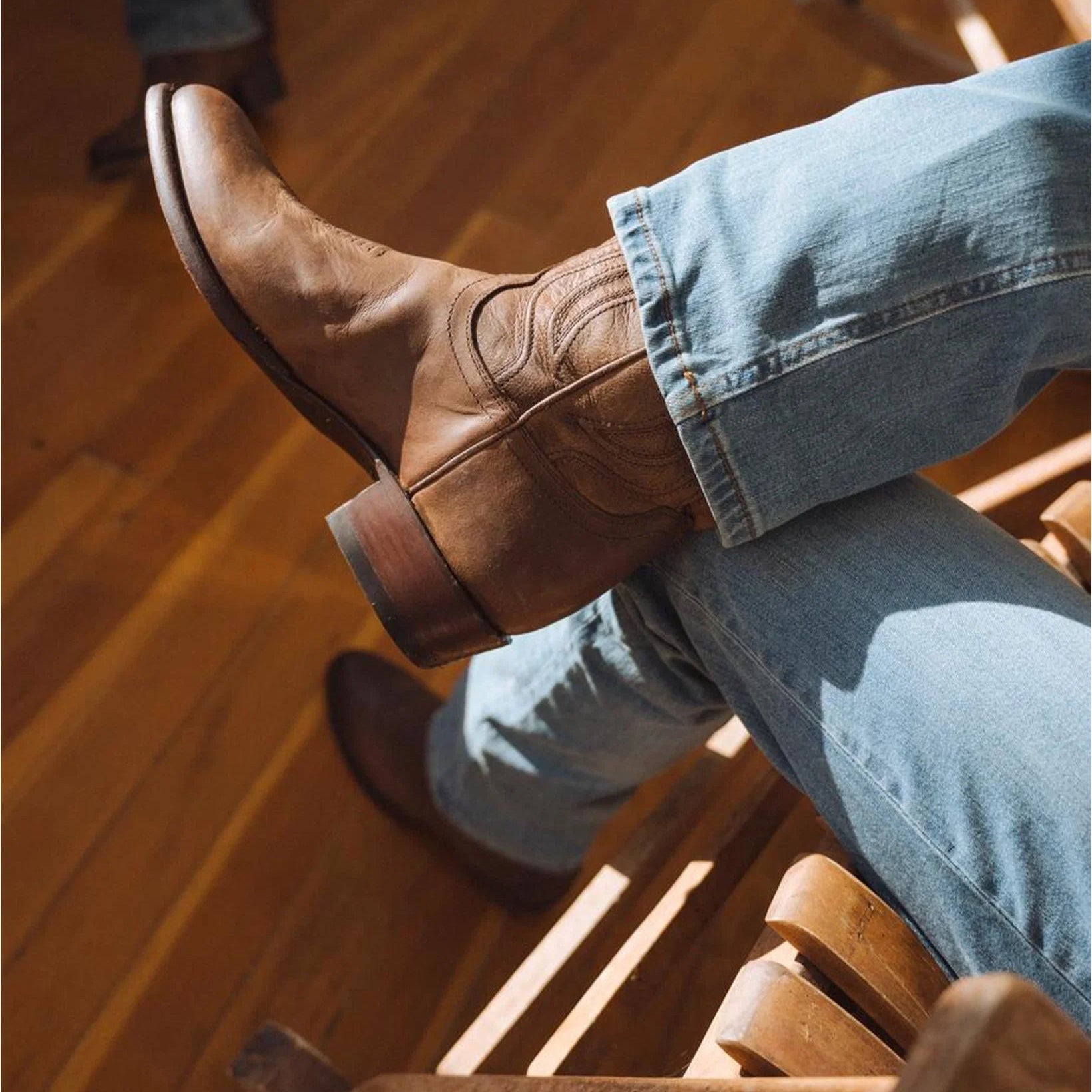 A person sitting on a wooden chair wearing a brown leather cowboy boot with western stitching, paired with light blue jeans, on a warm wood floor.
